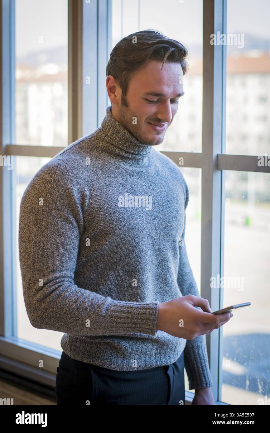 Hübscher trendigen Mann trägt Wolle Pullover stehen und blickte auf ein Handy, das er in der Hand, indoor inmitten der Stadt Stockfoto