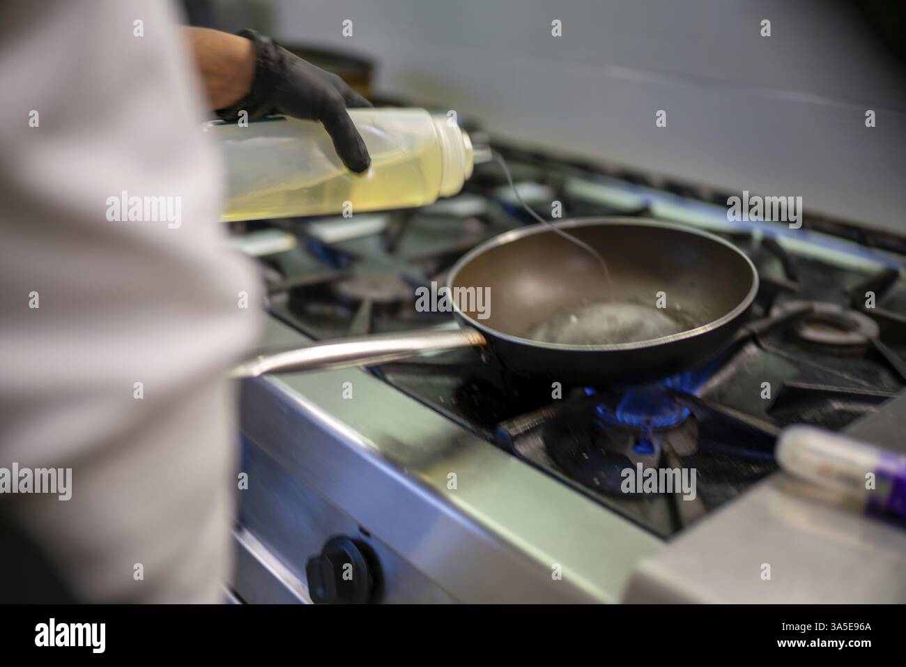 Der professionelle Koch trägt schwarze Handschuhe, gießt Öl aus einer Plastikflasche in eine Pfanne auf einem Gasherd und bereitet sich auf indische Gerichte in einem Restaurantset vor Stockfoto