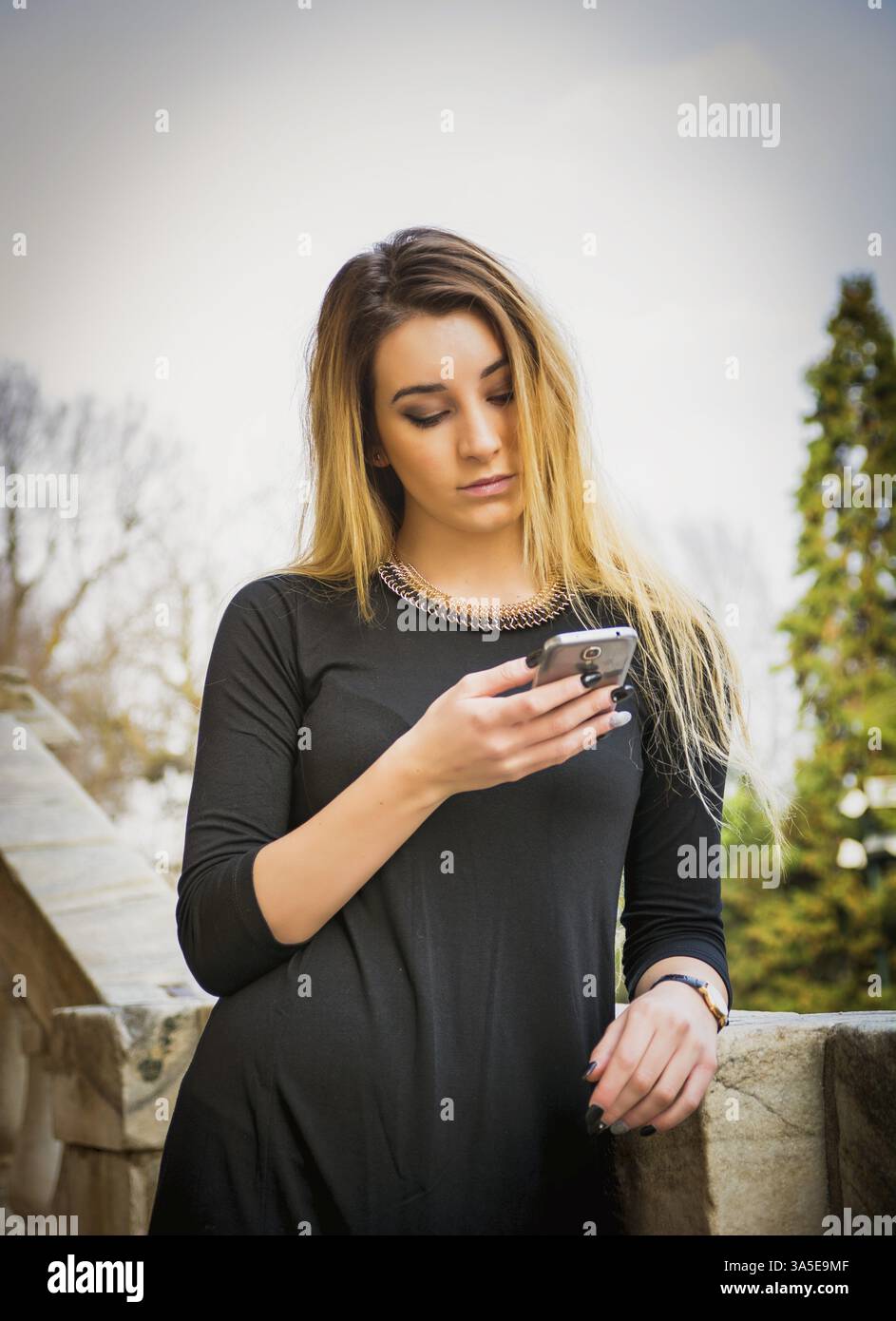 Schöne junge Frau mit langen Haaren, die auf dem Handy tippt und plaudert, während sie sich auf ein Steingeländer lehnt Stockfoto