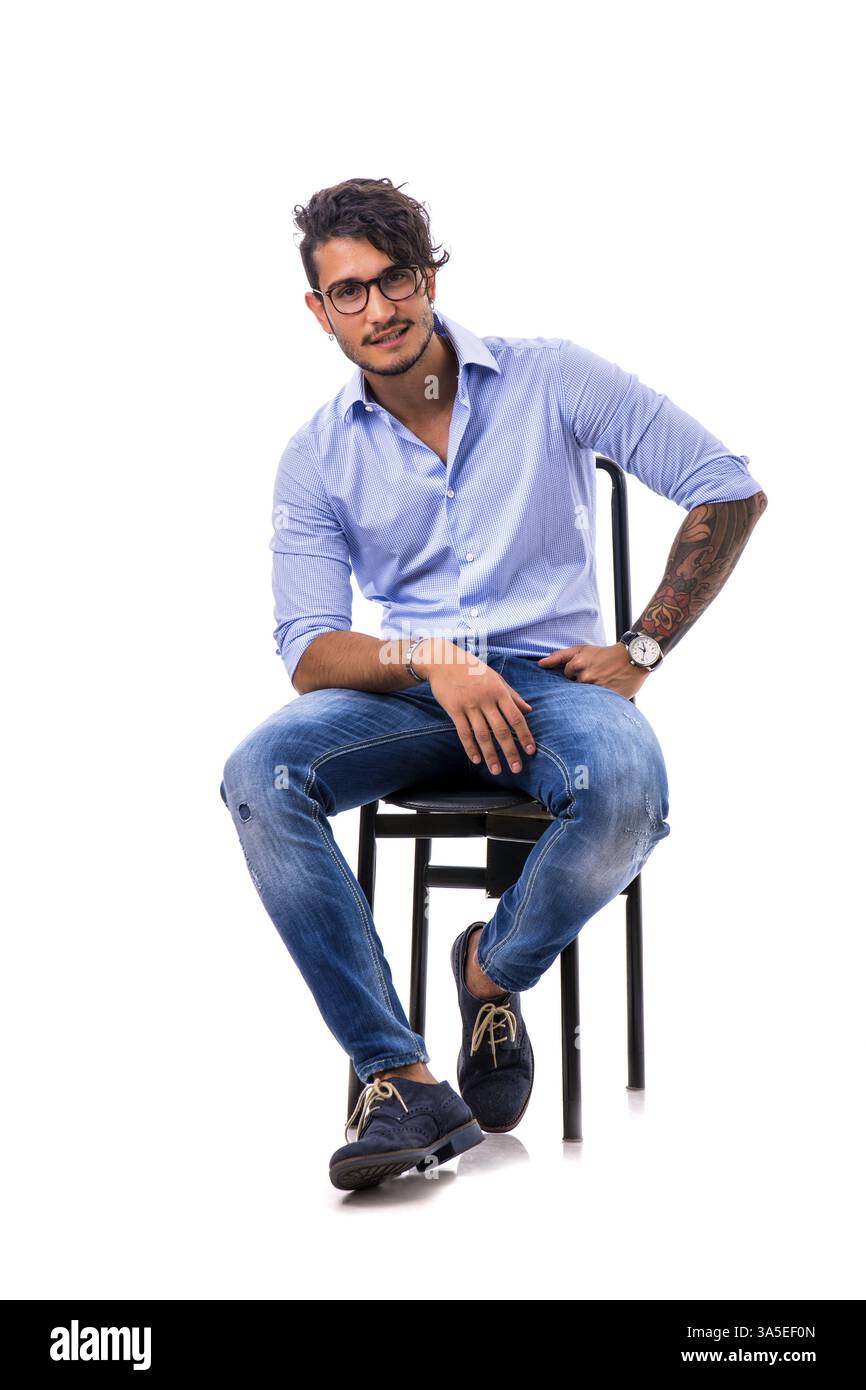 Porträt eines brünetten jungen Mannes in hellblauem Hemd und Jeans, sitzend auf Stuhl im Studio, isoliert vor weißem Hintergrund. Foto in voller Länge Stockfoto