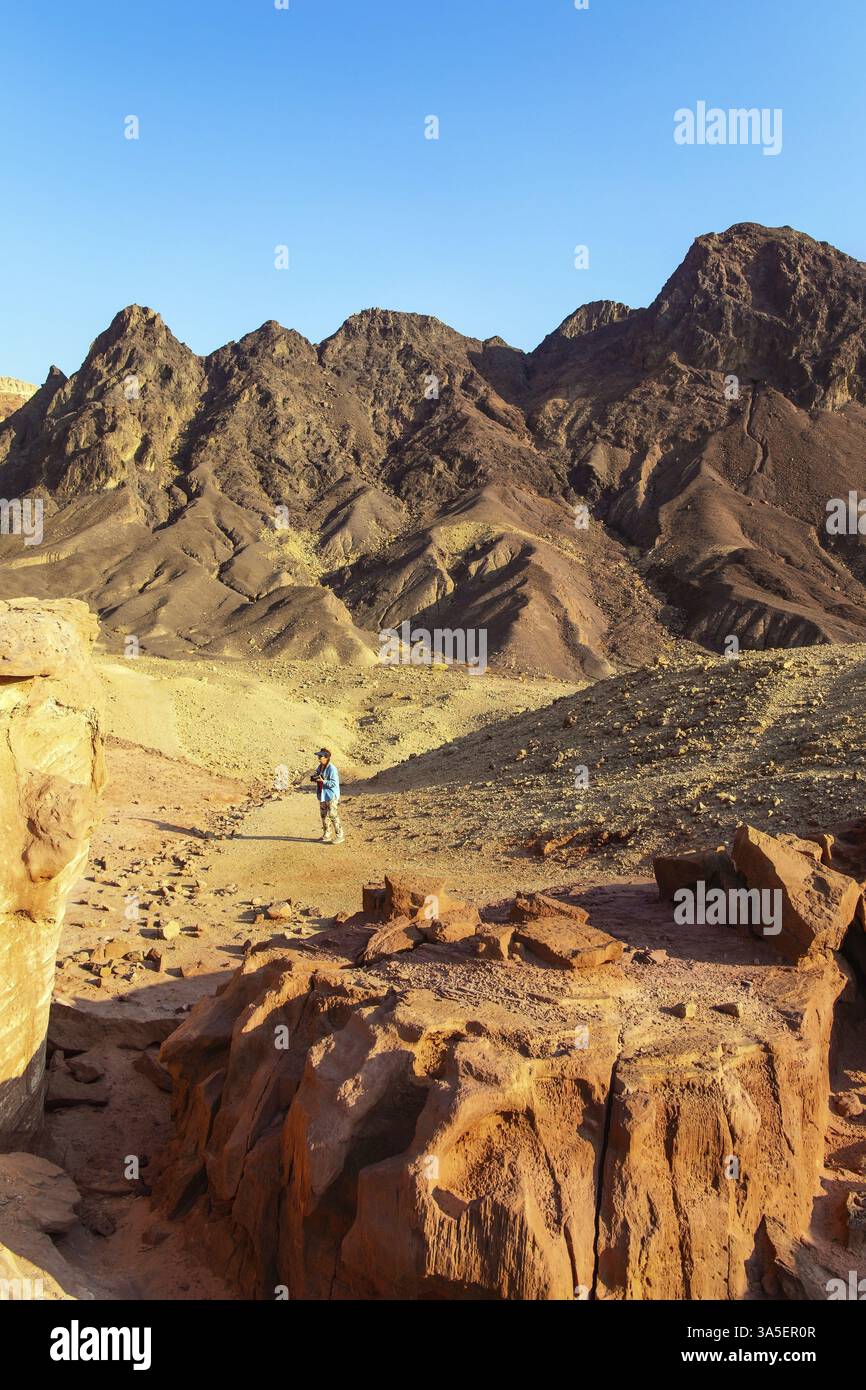 Frau in blauer Jacke fotografiert mit Begeisterung Felsen aus rot-orangenem Sandstein. Bunte Landschaftsformationen. Die Eilat Mountains. Israel Stockfoto