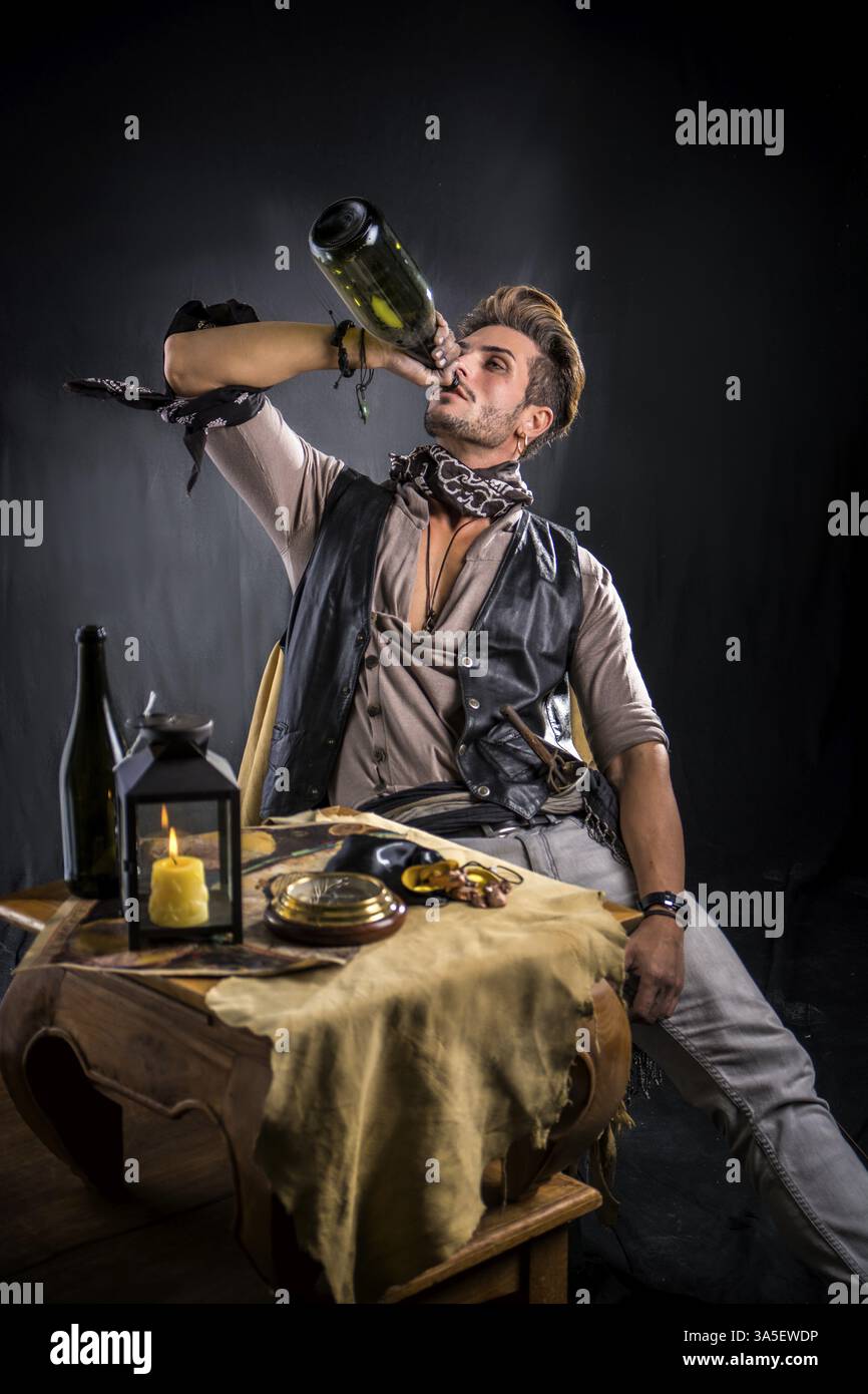 Gut aussehender junger Mann im Piratenmode-Outfit auf grauem Hintergrund. Gefangen im Studio, sitzen an einem Tisch mit einer Flasche Wein Stockfoto Gut aussehender junger Mann im Piratenmode-Outfit auf grauem Hintergrund. Gefangen im Studio, sitzen an einem Tisch mit einer Flasche Wein Stockfoto