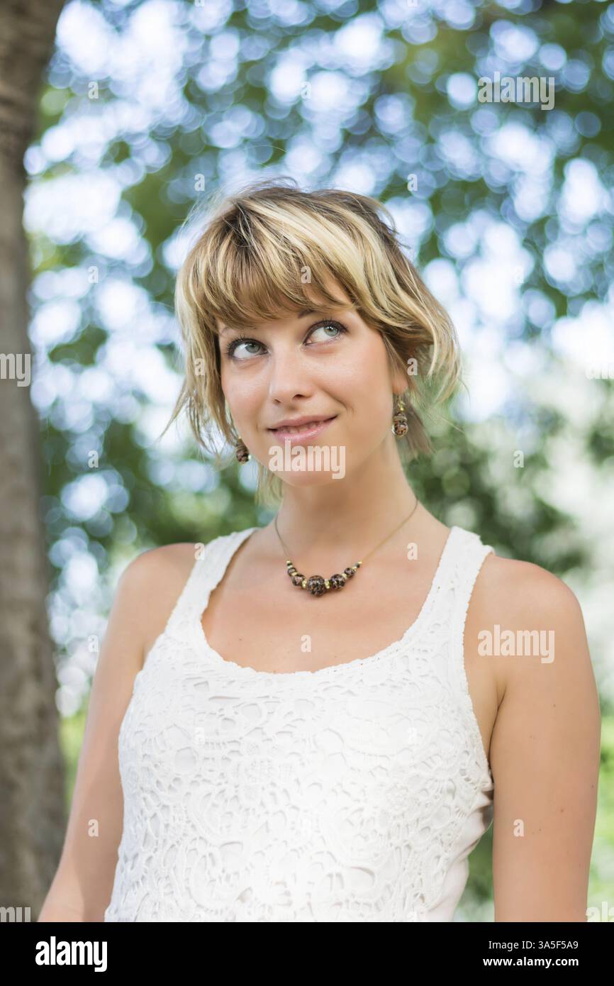 Attraktive junge Frau in einem stilvollen weißen Spitzentop im Garten, die tagträumt oder denkt und wünscht Stockfoto