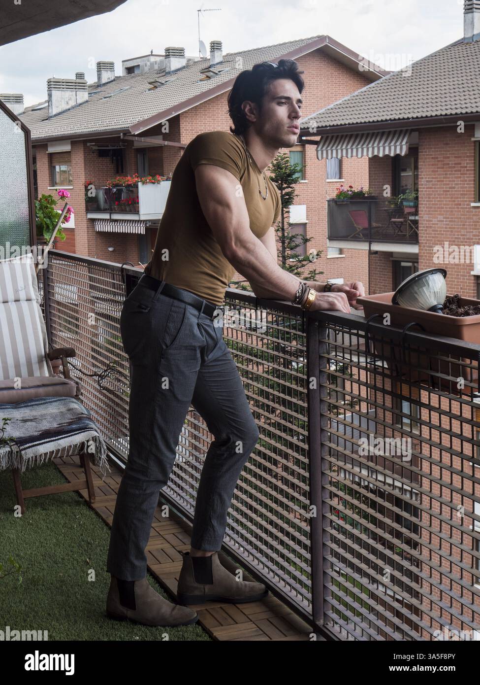 Hübscher dunkelhaariger junger Mann, der auf einem Balkon steht, aus- und runterblickt, mit braunem T-Shirt, im Sommer, Vollaufnahme Stockfoto