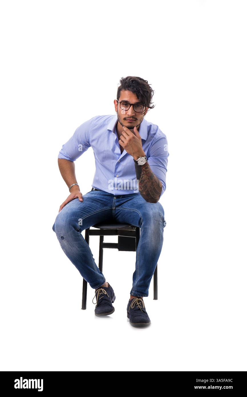 Porträt eines brünetten jungen Mannes in hellblauem Hemd und Jeans, sitzend auf Stuhl im Studio, isoliert vor weißem Hintergrund. Foto in voller Länge Stockfoto