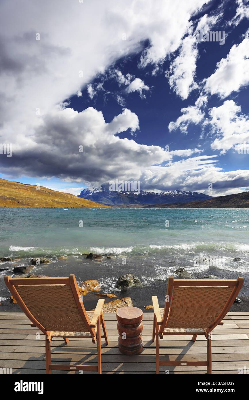 Zwei zusammenklappbare Holzstühle und ein kleiner Nachttisch auf der Promenade. Der komfortable Ort, um die Schönheit des Sees und der Wolken in Patagonien zu genießen Stockfoto