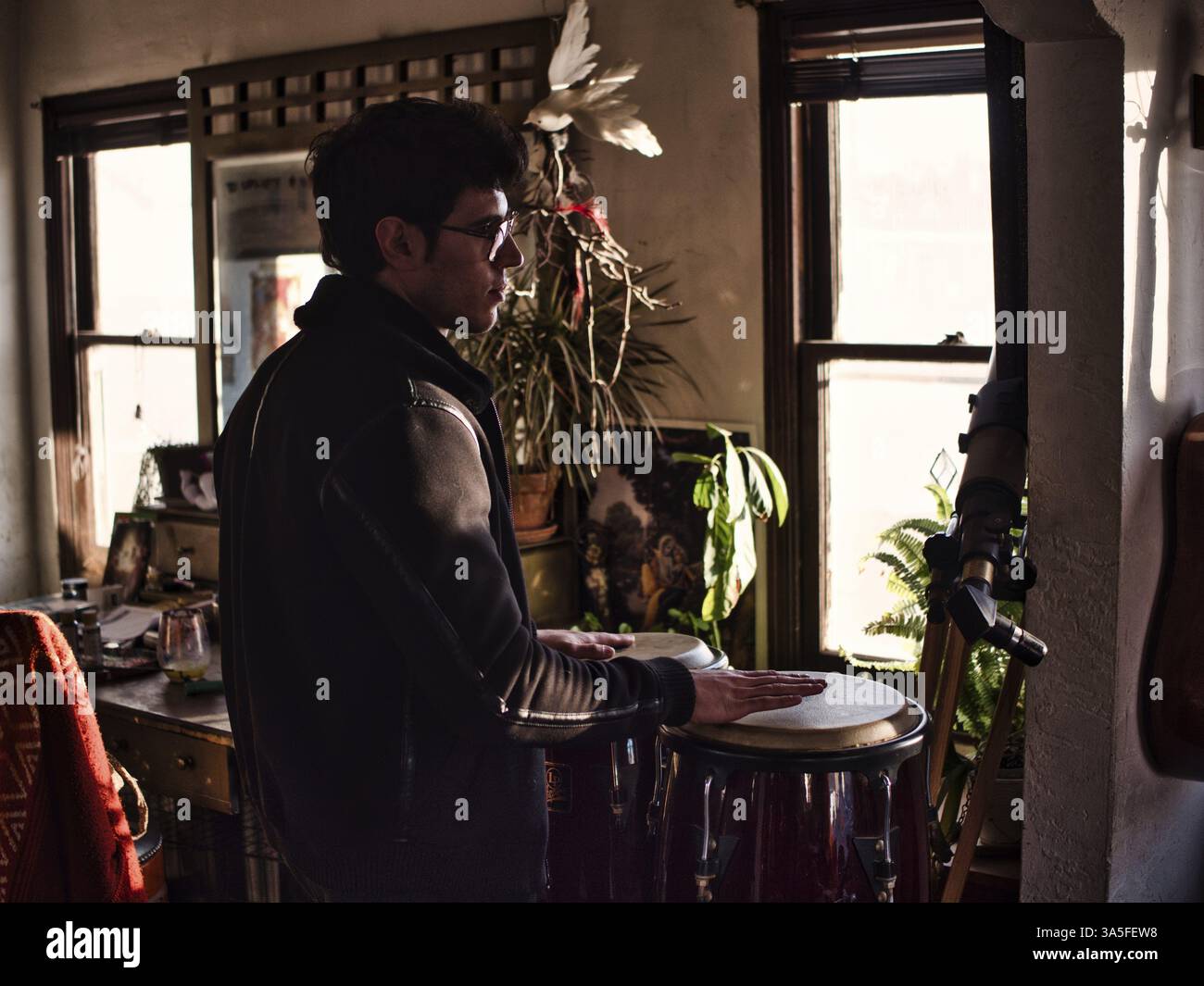 Seitenansicht des jungen Mannes in stilvoller Jacke und Brille Spielen Bongo Schlagzeug und Blick weg in gemütlichen Retro-Zimmer Stockfoto
