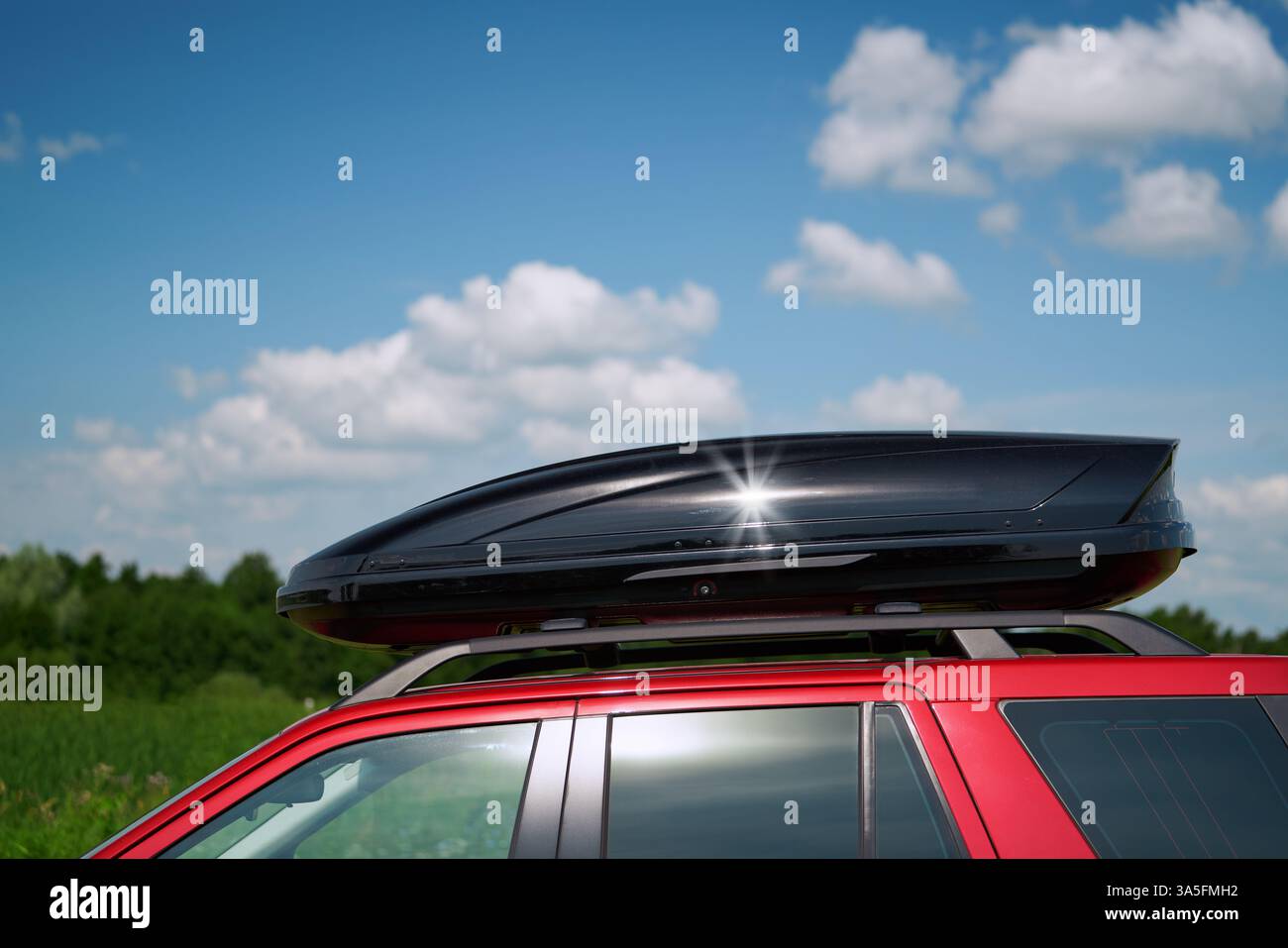 Nahaufnahme einer schwarzen Hochglanzdachbox an einem roten Auto. Im Hintergrund ist ein grüner Wald und ein blauer Himmel mit Wolken Stockfoto