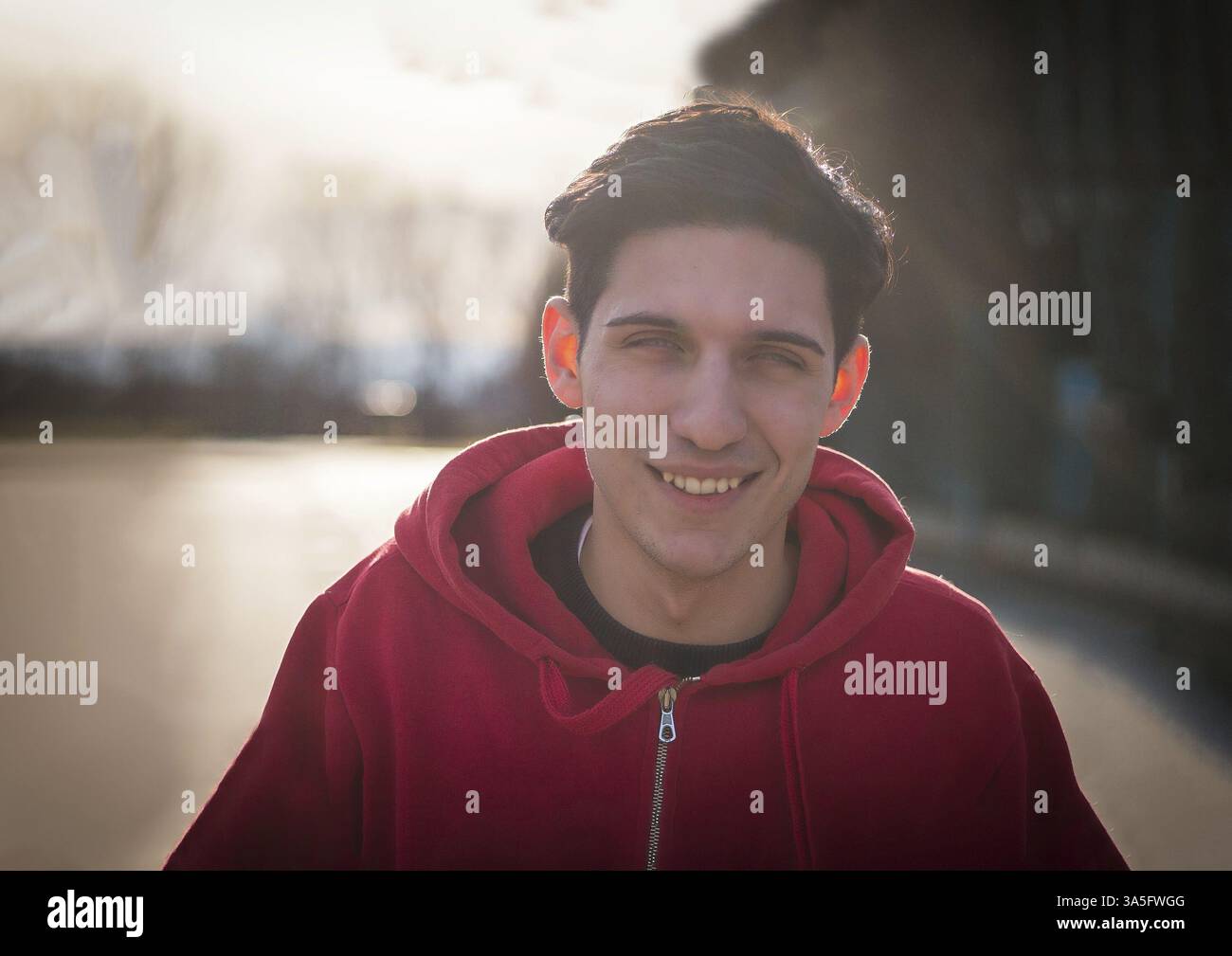 Ein junger Mann mit rotem Hoodie lächelt auf diesem Portraitfoto direkt an die Kamera. Sein Ausdruck ist fröhlich und einladend und strahlt ein Gefühl von fri aus Stockfoto