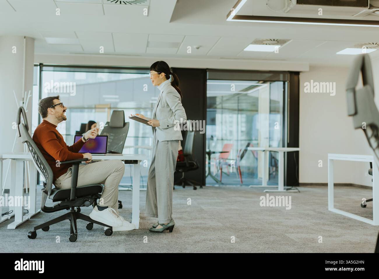 In einem modernen Büro arbeitet eine Frau in einem Anzug mit einem sitzenden Mann zusammen, während Kollegen an Schreibtischen in der Nähe arbeiten, um kreative Diskussionen und Teamarbeit zu fördern Stockfoto