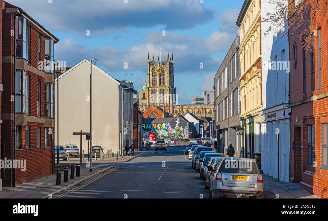 Eine Straße mit Ziegelsteinen und weiß getünchten Gebäuden säumt eine Stadtstraße mit Autos, die auf einer Seite geparkt sind. In der Nähe befindet sich eine Kathedrale mit einer Mauer Stockfoto Eine Straße mit Ziegelsteinen und weiß getünchten Gebäuden säumt eine Stadtstraße mit Autos, die auf einer Seite geparkt sind. In der Nähe befindet sich eine Kathedrale mit einer Mauer Stockfoto