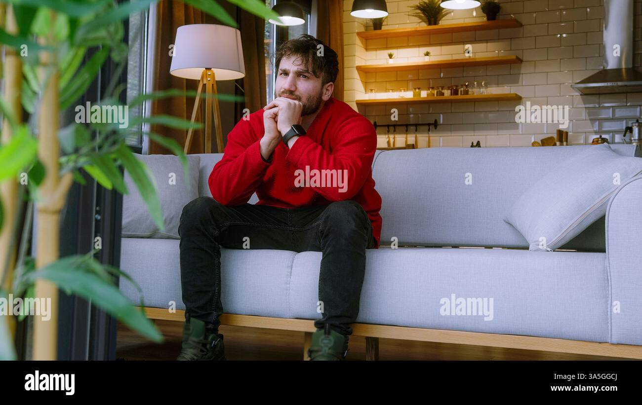 Ein nachdenklicher junger Mann, der auf einer gemütlichen Couch in einem modernen Haus sitzt, verloren in tiefem Denken Stockfoto