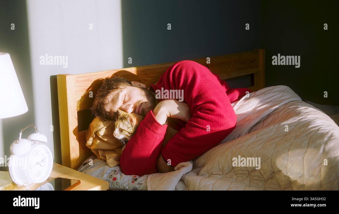 Mann schläft im Bett, während er seine Katze im warmen Morgensonnenlicht kuschelt – gemütlicher und friedlicher Moment der Begleitung und Entspannung mit Haustieren Stockfoto