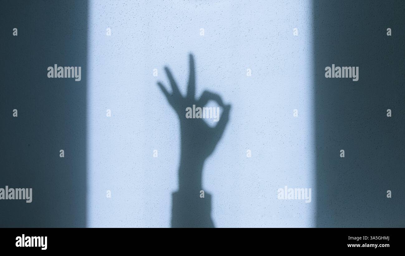 Der Schatten einer Hand, der die „OK“-Geste auf einer sonnendurchfluteten Wand macht, symbolisiert Zustimmung, Positivität und Kommunikation mit natürlichem Licht und starkem Kontrast Stockfoto