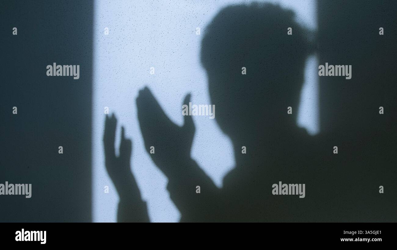 Schatten eines Mannes, der gegen eine sonnendurchflutete Wand betet, symbolisiert Glauben, Spiritualität und Hingabe mit starkem Kontrast und einem minimalistischen, kontemplativen Atmo Stockfoto