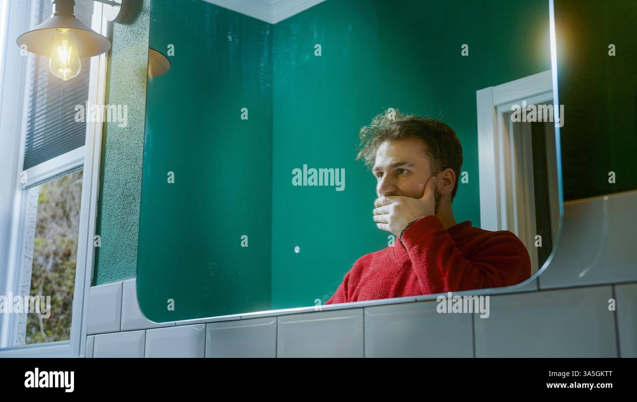 Männlicher Mann in einem roten Pullover, der vor einem Spiegel steht und seine Ohren bedeckt, als Teil des Konzepts "kein Böses sehen, kein Böses hören, kein Böses sprechen", expre Stockfoto