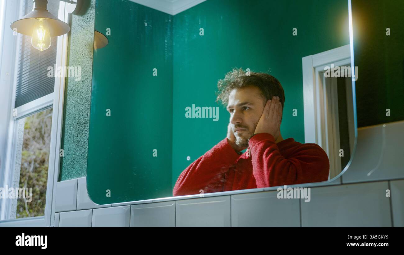 Junger Mann in einem roten Pullover, der vor einem Spiegel steht und seine Ohren bedeckt, als Teil des Konzepts "kein Böses sehen, kein Böses hören, kein Böses sprechen", ausgedrückt Stockfoto