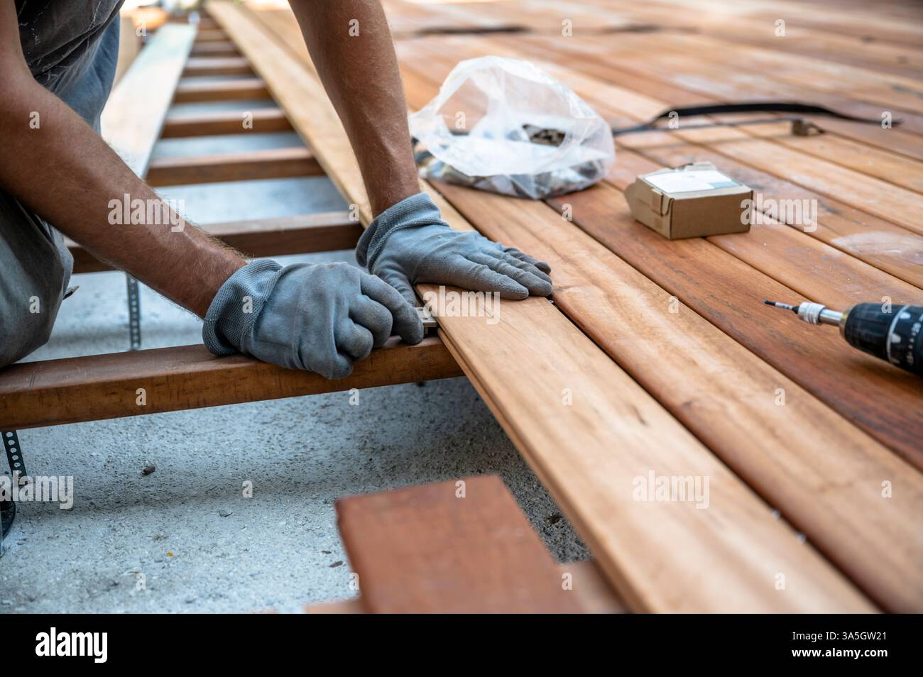 Ein Arbeiter, der Handschuhe trägt, richtet beim Bau eines Decks sorgfältig Holzdielen aus, wobei Werkzeuge und Materialien wie Bohrer und Schrauben auf der sur sichtbar sind Stockfoto