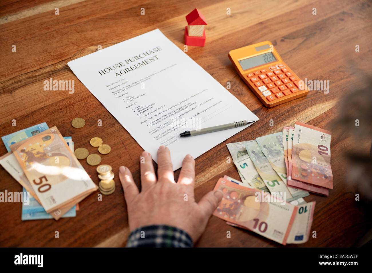 Eine Hand liegt auf einem Hauskaufvertrag, umgeben von Euro-Banknoten, Münzen, einem Taschenrechner und einem kleinen roten Hausmodell auf einem Holztisch. Stockfoto