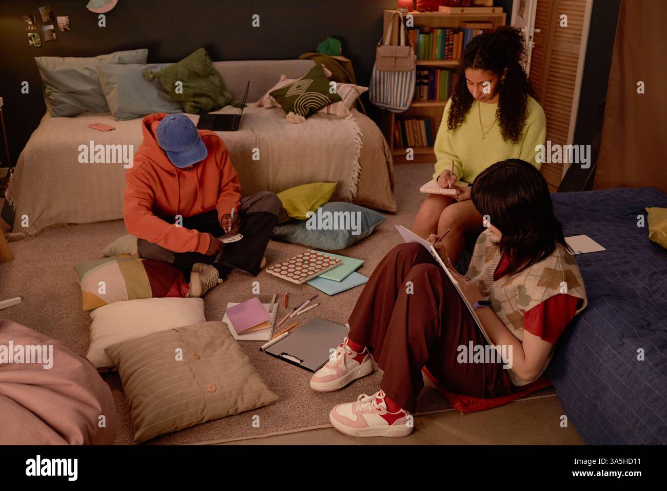 Gruppe aus drei verschiedenen Studenten, die sich auf das Studium in einem gemütlichen Wohnzimmer mit Sitzgelegenheiten, Büchern und Notizbüchern konzentrieren. Jeder Schüler konzentrierte sich auf die individuelle Aufgabe und setzte sich bequem im Sitzen Stockfoto