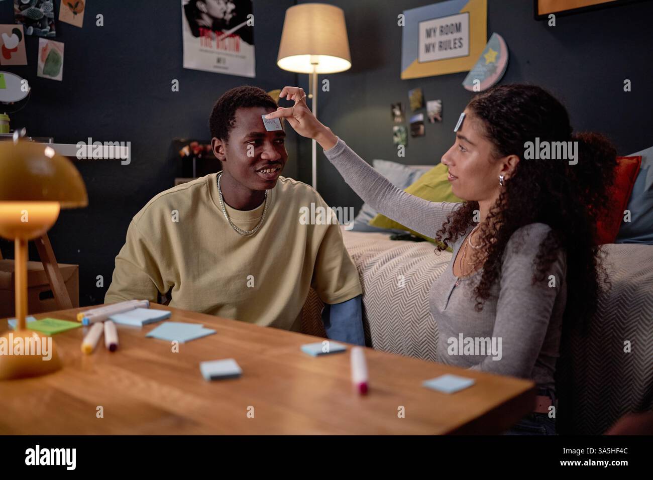 Junger Mann mit fröhlichem Ausdruck, der mit einer Frau Ratespiele in einem Wohnzimmer voller warmer Beleuchtung und persönlicher Einrichtung spielt. Sie verwenden Haftnotizen auf ihrer Stirn Stockfoto