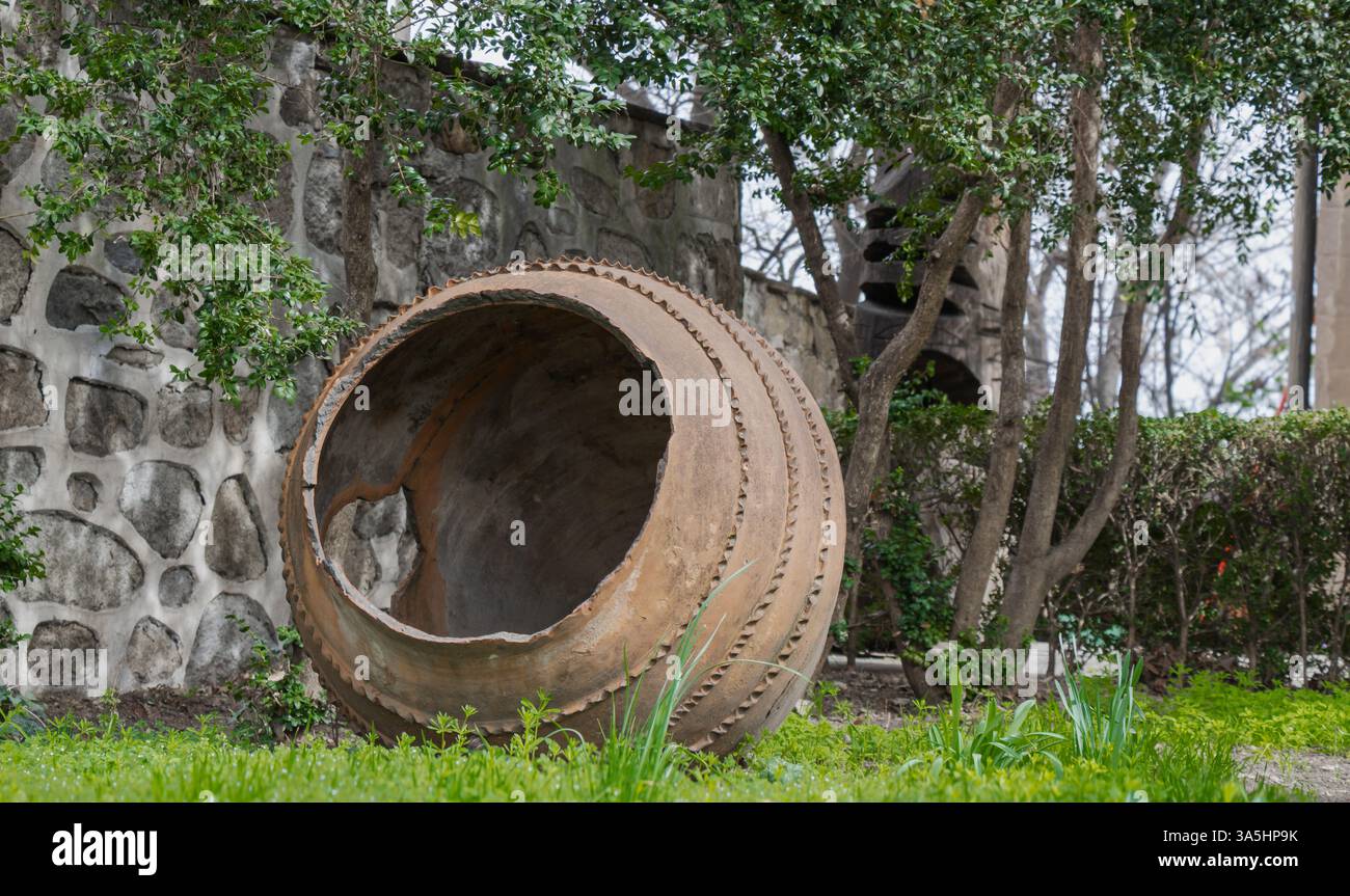Ein übergroßer Tontopf liegt gegen eine verwitterte Steinmauer, umgeben von lebendigem Grün. Die ruhige Gartenanlage lädt zum Nachdenken ein Stockfoto
