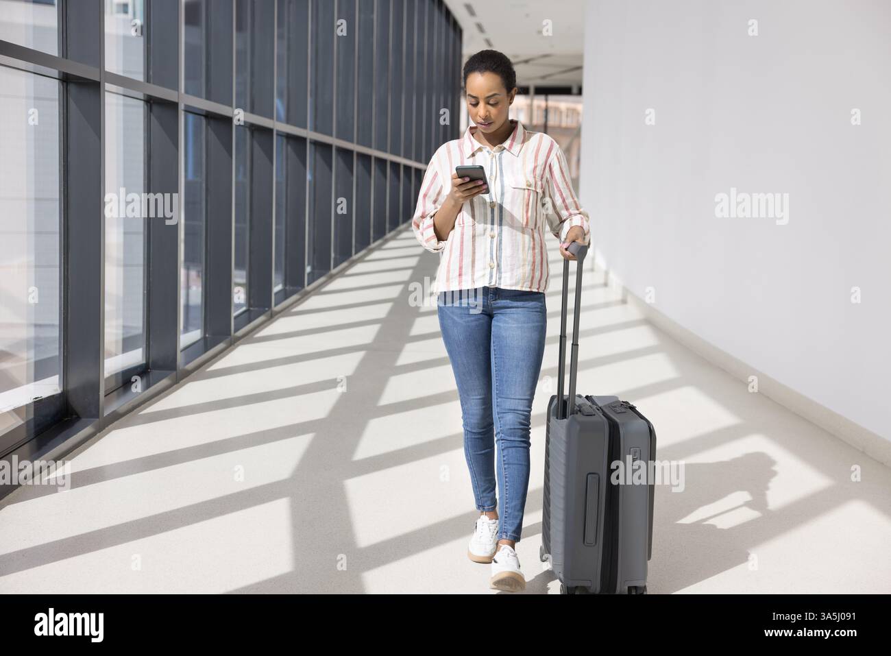Afrikanische Reisende Frau, die lässig gekleidet ist, durch einen modernen Flur mit großen Panoramafenstern, rollendem Koffer, mit Smartphone unterwegs Stockfoto