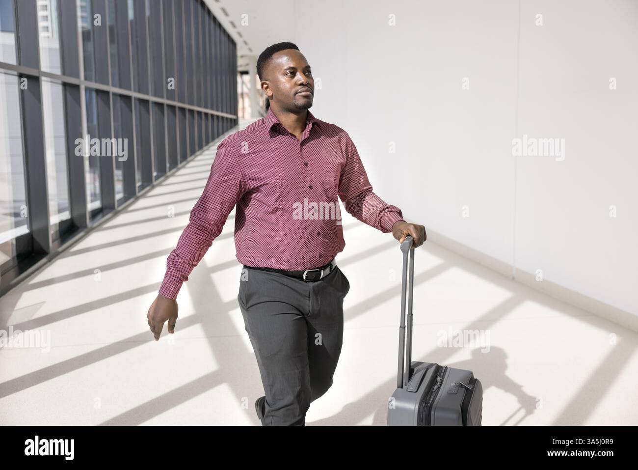 Junger afrikanischer Geschäftsmann, der zu einer Arbeitskonferenz oder einem Geschäftstreffen in eine andere Stadt reist, seinen Koffer Rollen und sich selbstbewusst in Richtung Gatter bewegt Stockfoto