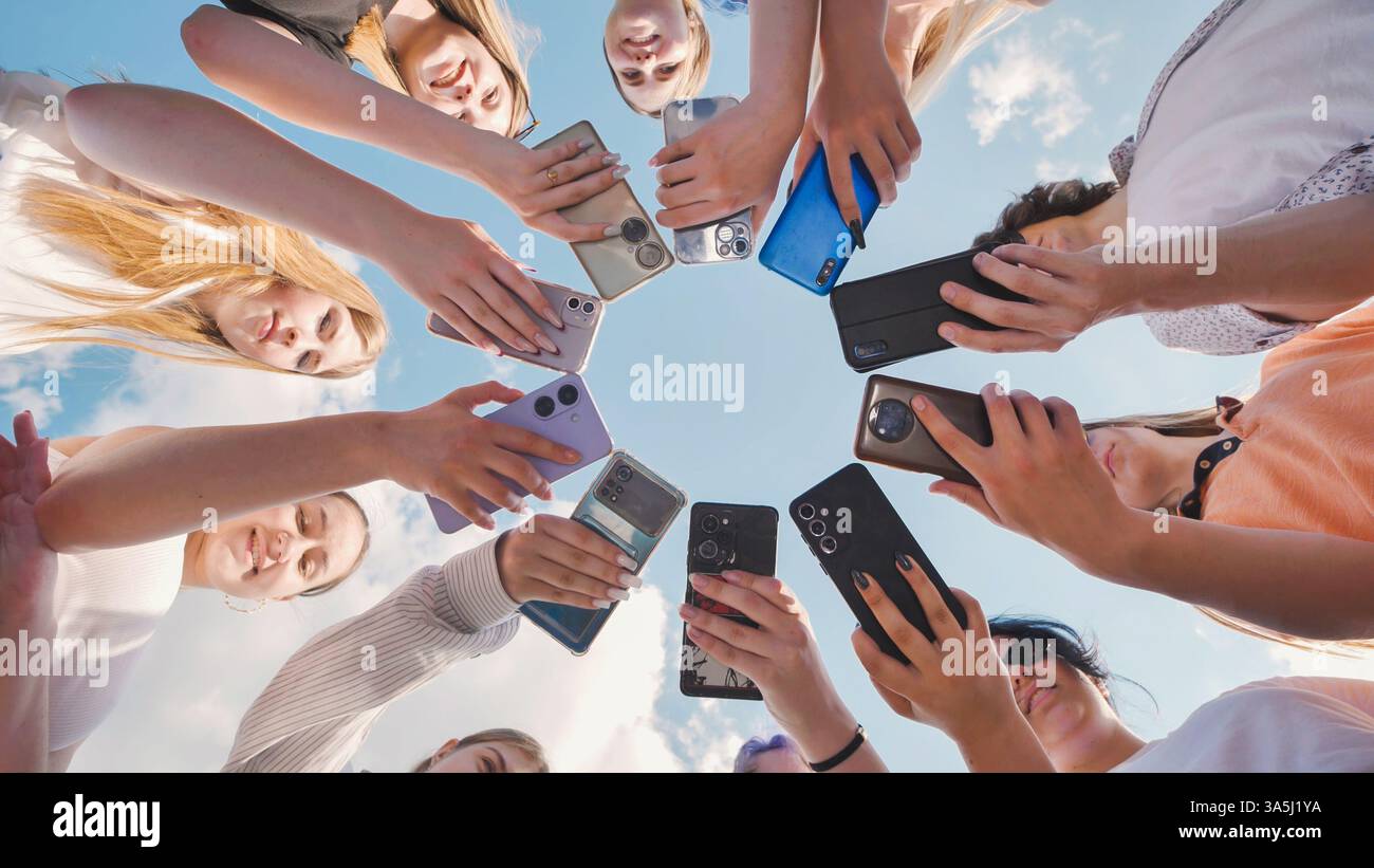 Schulfreunde treffen sich im Kreis, verwenden Smartphones, um Inhalte zu teilen und gemeinsam Spaß im Freien zu haben Stockfoto