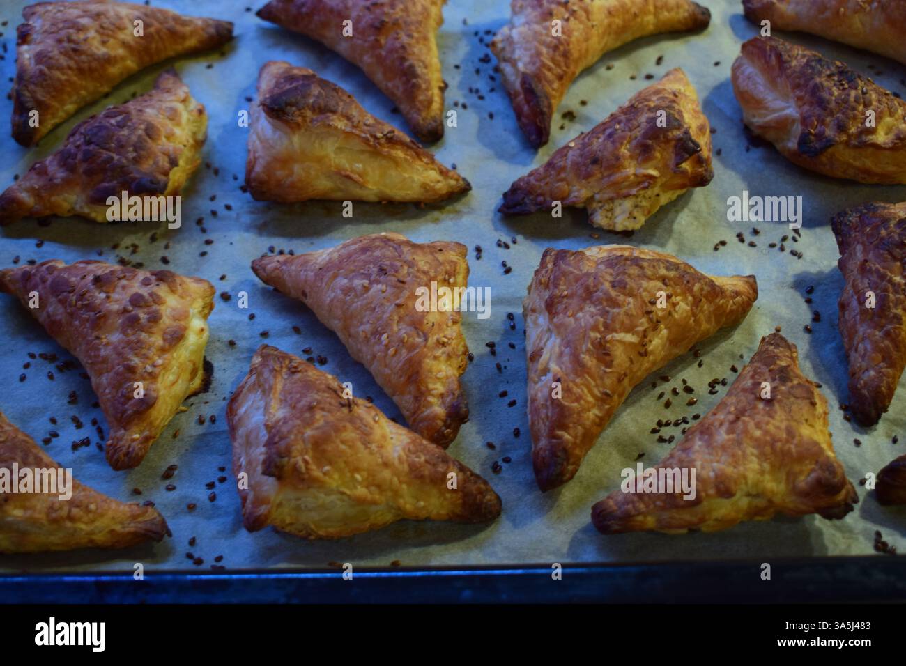Ein Backblech gefüllt mit frisch gebackenen goldbraunen dreieckigen Kuchen. Die Kuchen haben eine blättrige, knusprige Textur und sind mit Sesam bestreut. Stockfoto