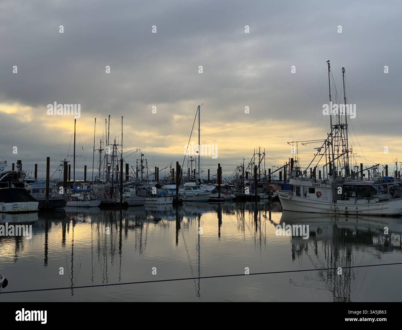 Friedlicher Hafen auf dem Meer, Fischerboote ruhen in ruhigen Gewässern unter einem weichen Sonnenuntergang Himmel. Warme goldene Sonnenlichttöne verleihen Reflexen ein warmes Leuchten. - Smartphone-aufgenommenes Stockfoto