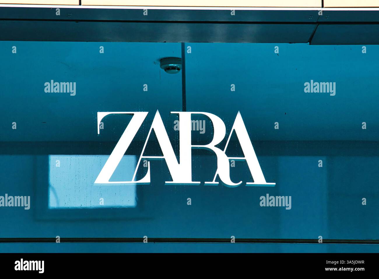 Eine Nahaufnahme des ZARA-Logos, weiß, auf einer Ladenfront aus blauem Glas mit Reflexionen und einer Sicherheitskamera, sichtbar in Manchester, Großbritannien. Stockfoto
