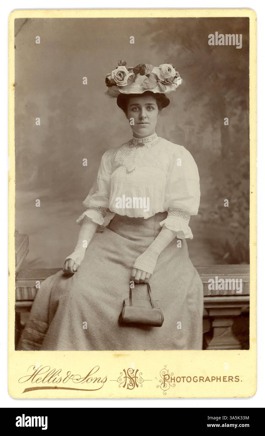 Originale edwardianische Schrankkarte der hübschen edwardianischen Frau, edwardianischen Dame, die Mode im Frühling oder Sommer trägt - eine weiße Bluse aus Leinen und Spitze, weiße Handschuhe aus Spitze, ein Hut mit Rosen, eine kleine Handtasche in der Hand, Studio of Hellis & Sons Photographers, London, datiert vom Juli 1907 auf der Rückseite. Stockfoto