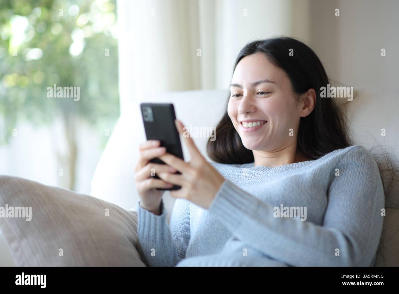 Glückliche asiatische Frau benutzt ihr Smartphone und sitzt zu Hause auf einer Couch Stockfoto