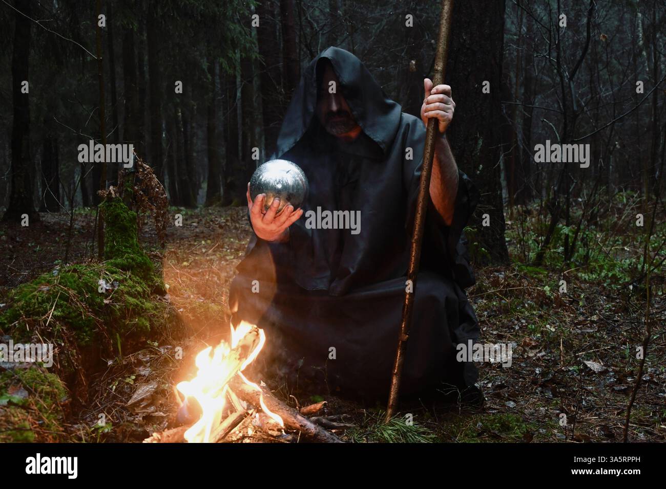 Ein Mönch in schwarzem Gewand sitzt am Feuer im Wald und schaut in einen Wahrsagungsball. Mystik und Magie, Zauberer und Hexen. Stockfoto