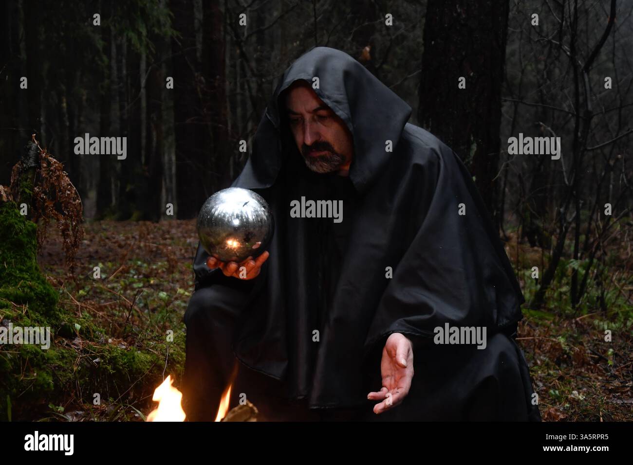 Ein Mönch in schwarzem Gewand sitzt am Feuer im Wald und schaut in einen Wahrsagungsball. Mystik und Magie, Zauberer und Hexen Stockfoto