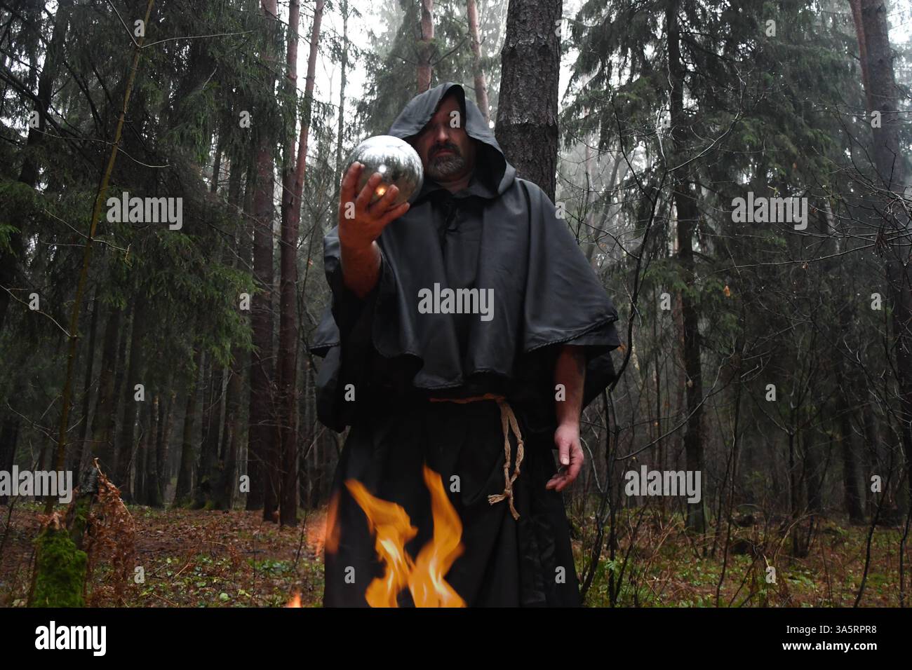 Ein Mönch in schwarzem Gewand sitzt am Feuer im Wald und schaut in einen Wahrsagungsball. Mystik und Magie, Zauberer und Hexen. Stockfoto