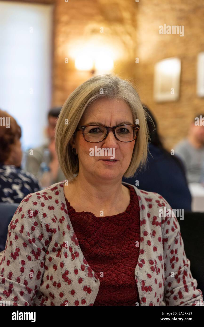 Ein Porträt einer Frau mittleren Alters, die drinnen sitzt. Sie ist umgeben von Menschen in einer warmen sozialen Umgebung, die eine freundliche und einladende Atmosphäre schafft Stockfoto