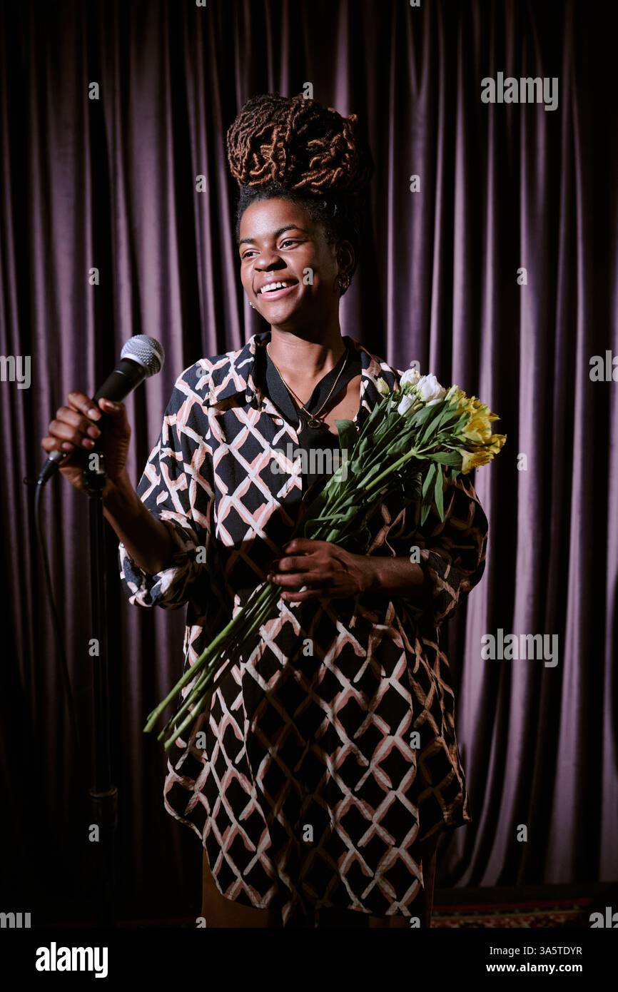 Frau mit geflochtenen Haaren lächelt, während sie das Mikrofon hält und Blumenstrauß auf der Bühne mit Vorhangfassade hält. Mit brillantem Ausdruck aufführen oder präsentieren Stockfoto