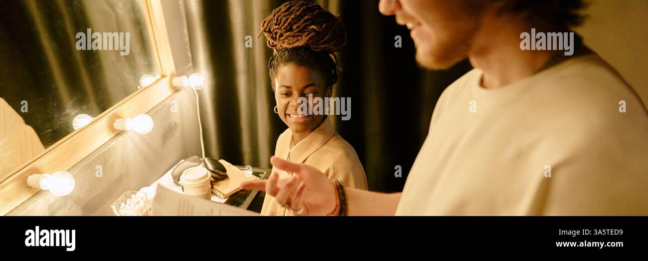 Afroamerikanische Frau mit Zöpfen, die lächelnd im Ankleidezimmer mit hellen Lichtern und Spiegeln zuhören Stockfoto