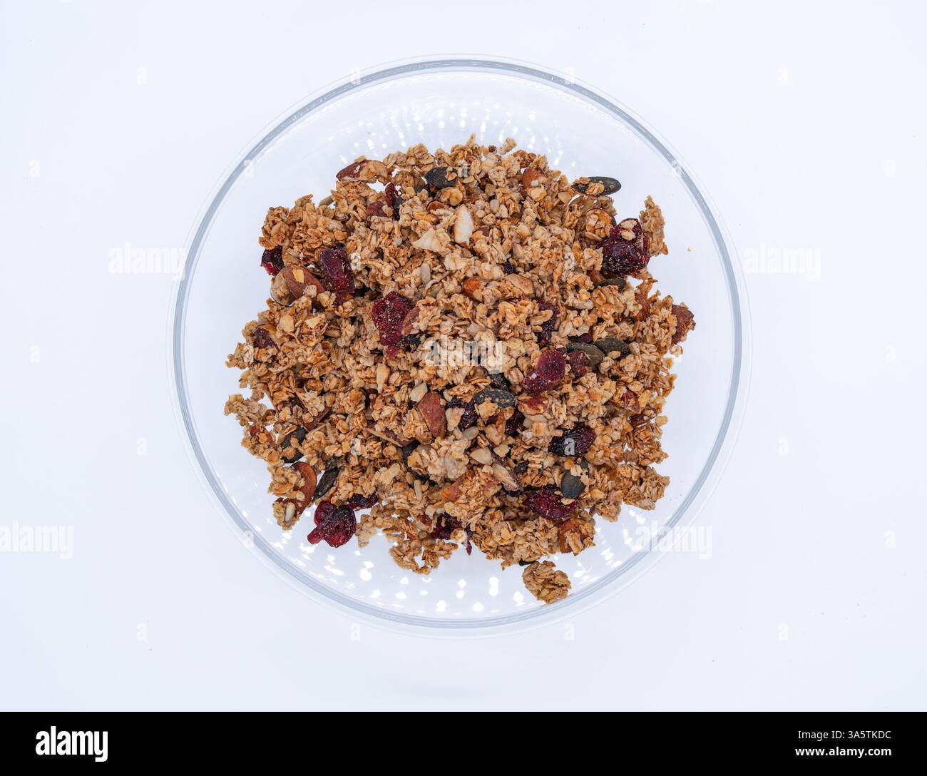 Eine klare Glasschale gefüllt mit Müsli, mit einer Mischung aus Hafer, Nüssen und getrockneten Früchten, einschließlich Preiselbeeren und Kürbiskernen, auf einem weißen Hintergrund Stockfoto