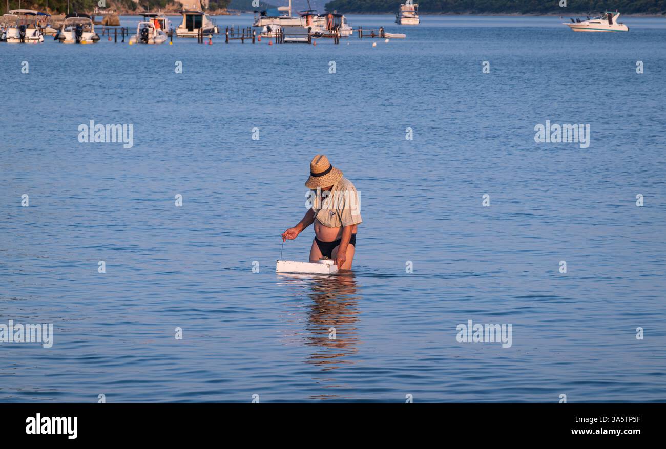 Rab, Kroatien - 24. Juli 2024: Eine Person, die in flachem Wasser steht, einen Strohhut und leichte Kleidung trägt und an einer Aktivität teilnimmt. Der Hintergrund Stockfoto