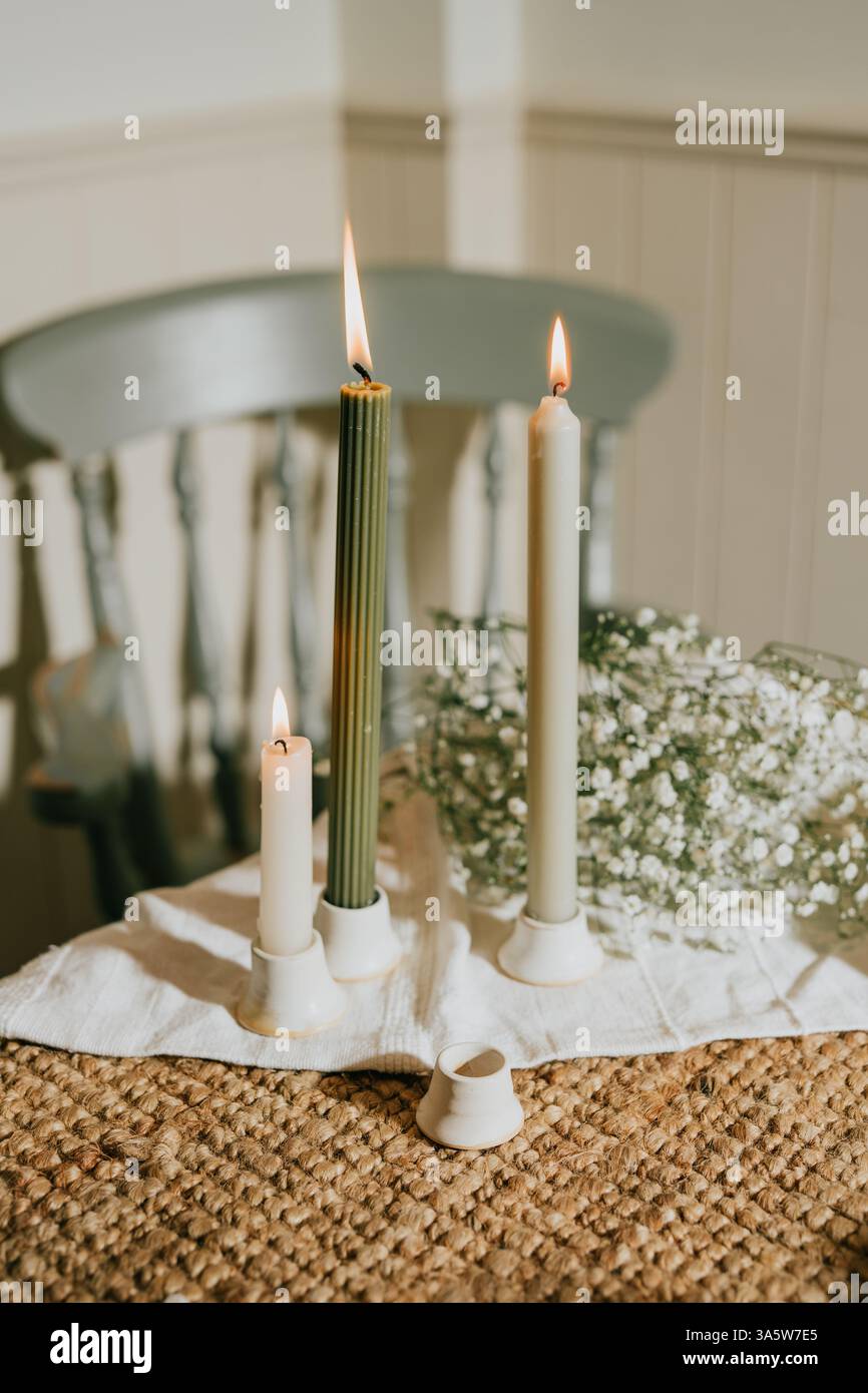 Ein Tisch mit drei Kerzen und einer Vase mit Blumen. Die Kerzen sind angezündet und die Blumen sind weiß. Die Szene ist warm und einladend Stockfotografie