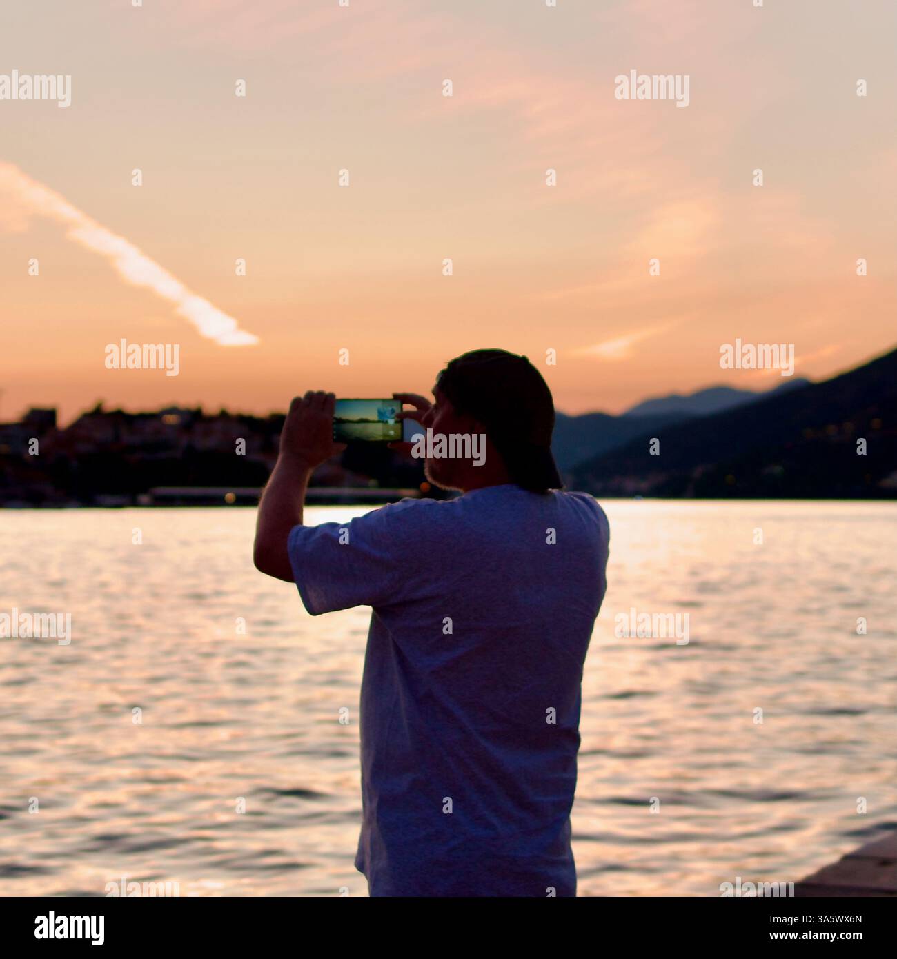 Eine Person fängt den ruhigen Sonnenuntergang über einem ruhigen See mit einem Smartphone ein, mit Bergen und orangefarbenem Himmel als Kulisse, die Frieden ausdrücken Stockfoto