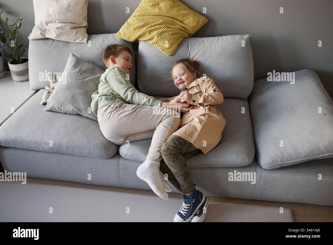 Porträt eines niedlichen Mädchens mit Down-Syndrom, das mit Bruder spielt und glücklich lacht, während er auf der Couch, im Kopierraum spielt Stockfoto