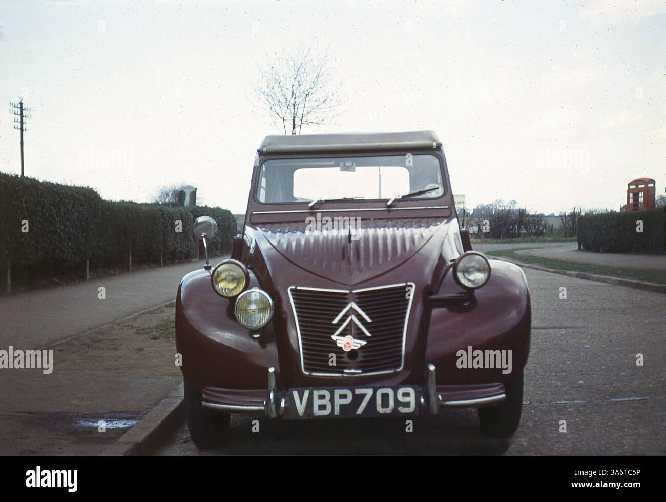 1966, historisch, ein Citroen 2CV, Vorderansicht, mit britischem Nummernschild, Burgund oder weinrot in Farbe, geparkt auf der Straße in England, Großbritannien. Eine britische rote Telefonzelle an der Straßenecke. Citroen, ein führender französischer Automobilhersteller, hatte in dieser Zeit eine Fabrik in Slough, England, die seine Autos für den britischen Markt herstellte. In der Produktion von 1948 bis 1990 gilt der Citroen 2CV als Designklassiker. Der Name leitet sich von „zwei Pferden“ ab... wie in „zwei steuerpflichtige Pferdestärken“ Stockfoto
