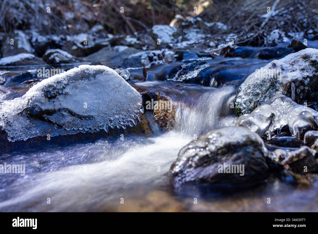 Kristallklares Wasser fließt über Felsen, die mit Schnee und Eis bedeckt sind, und schafft eine malerische Winterszene in einem Gebirgsbach Stockfoto