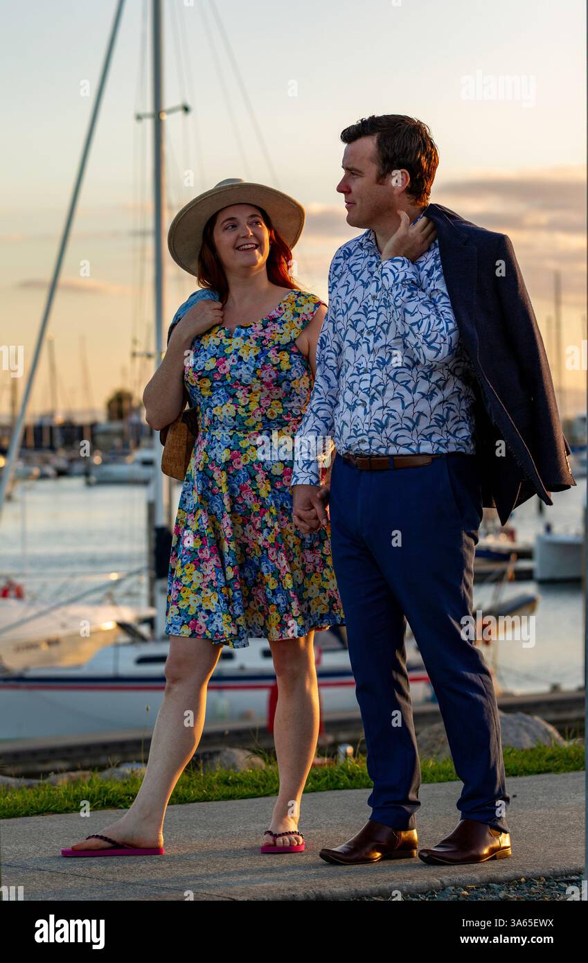 Ein elegantes Paar genießt einen Spaziergang in der Nähe eines malerischen Yachthafens bei Sonnenuntergang, trägt stilvolle, lebendige Sommerkleidung, die ruhige Atmosphäre und das malerische bac Stockfoto