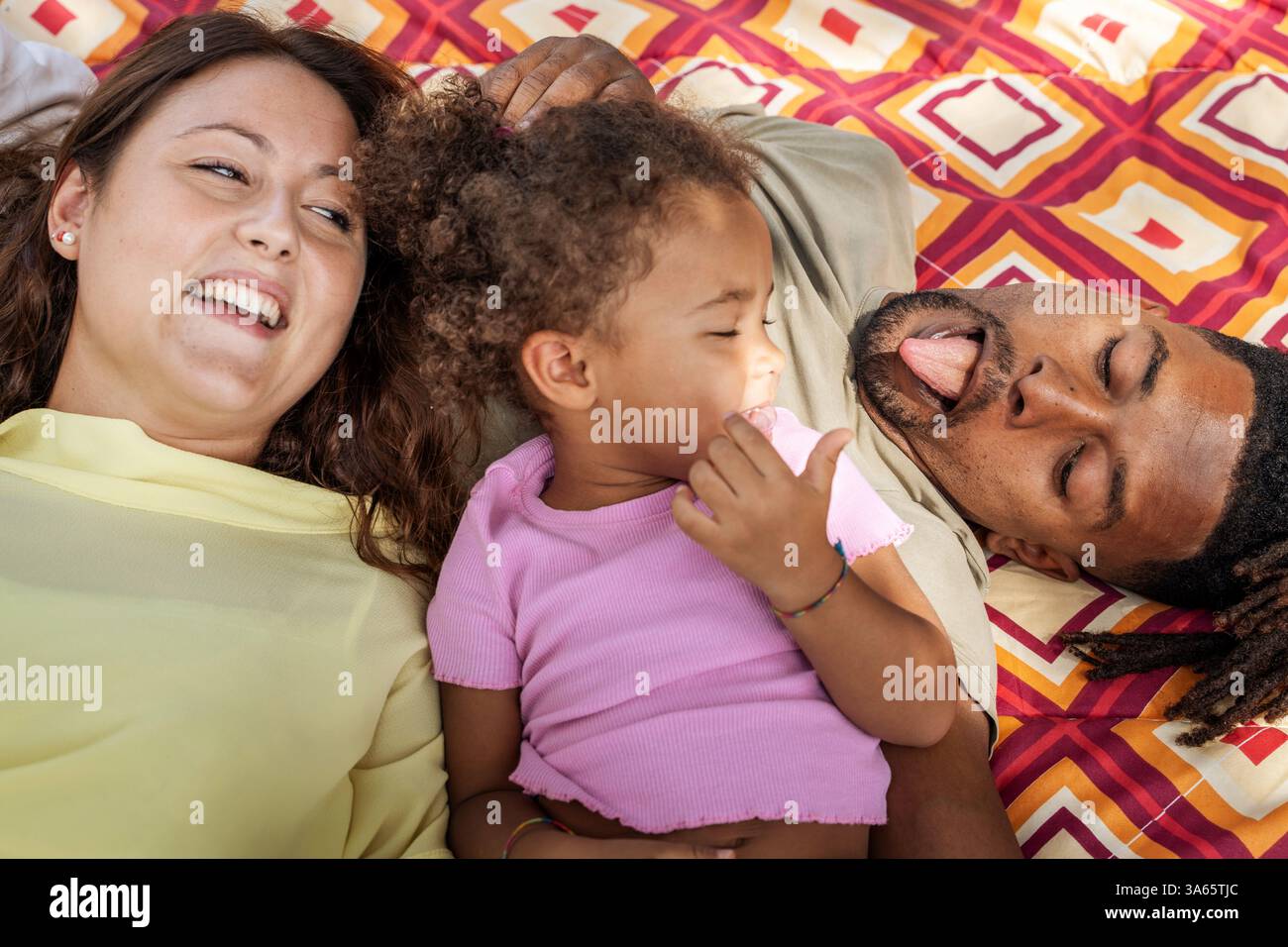 Lachende, vielfältige Familie, die sich auf einer gemusterten Picknickdecke im Park entspannt und einen verspielten Moment im Freien genießt. Stockfoto