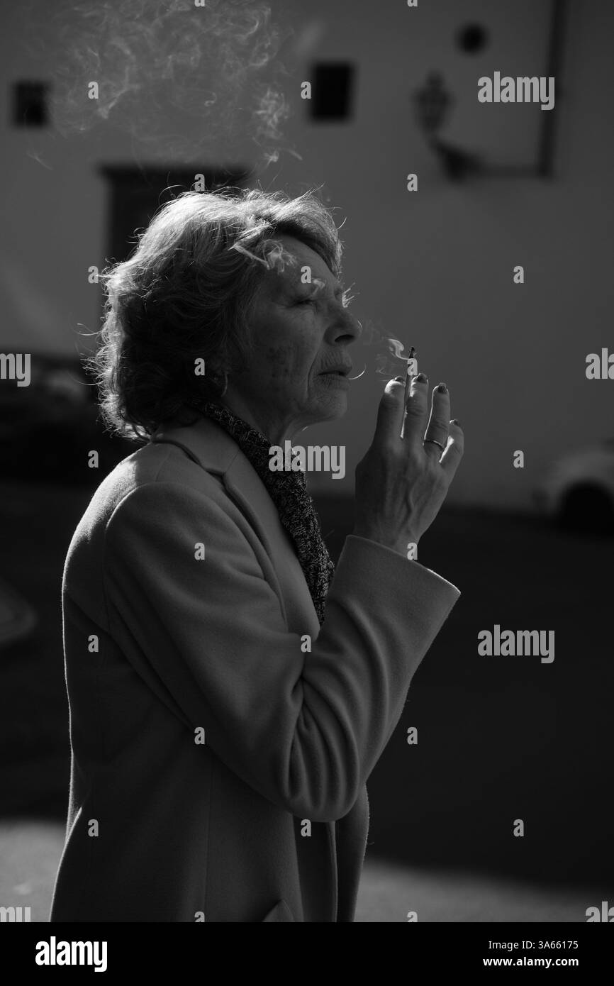 Eine ältere Frau steht draußen, hält eine Zigarette in der Hand, in Gedanken. Rauch weht sanft in die Luft, erfasst in auffälligem Schwarz-weiß und zeigt Stockfoto
